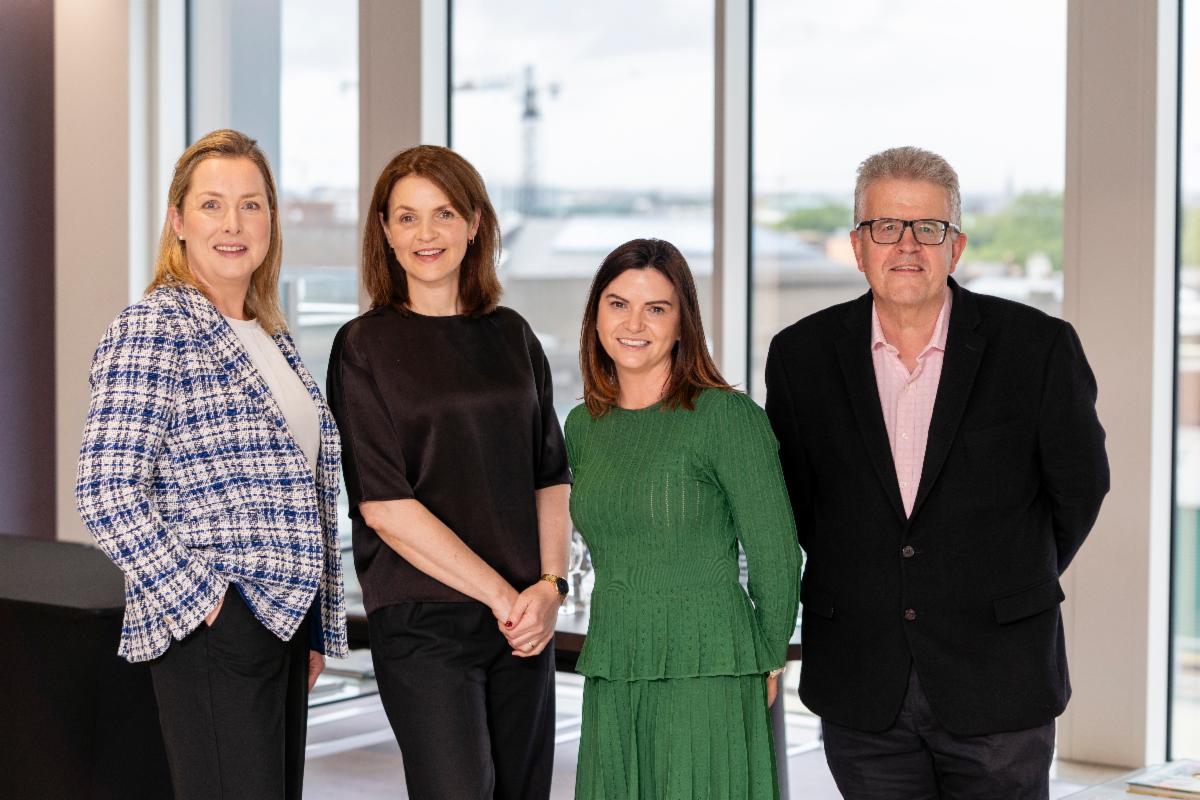 four people standing indoors in front of large windows with a cityscape view in the background the individuals have their faces obscured and are dressed in business casual attire