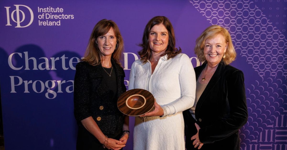 Three individuals pose in front of a purple Institute of Directors Ireland Chartered Director Programme Awards backdrop. The individual in the centre holds a wooden award plaque, while the two individuals on either side stand alongside. The IoD Ireland logo and Chartered Director Programme Awards branding are visible in the background.