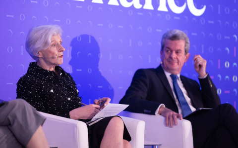 Two individuals seated on white chairs on a stage with a purple backdrop featuring text and patterns. One person is holding papers and wearing a dark outfit with decorative details. The other person is dressed in a dark suit with a white shirt and tie resting one hand near the face. A third person’s arm and leg are partially visible on the left side of the image.