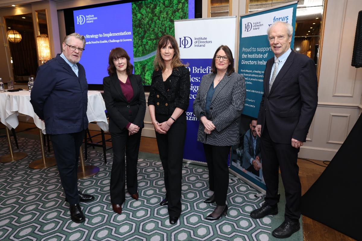 Group of five business professionals standing together at an Institute of Directors Ireland event venue. They are positioned in front of branded presentation screens and sponsor banners in a formal indoor setting with patterned flooring and soft lighting.