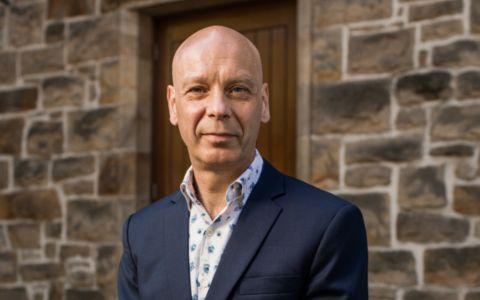 Professional portrait of a man wearing a navy blazer over a patterned shirt. He is standing outdoors in front of a stone wall and wooden doorwa