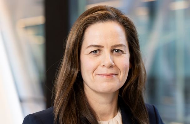 person with long brown hair wearing a dark blazer and light colored blouse standing in front of a blurred background with glass windows and structures