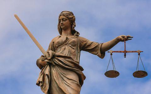 The image shows a statue of Lady Justice holding a sword in the left hand and a set of scales in the right hand. The face of the statue is covered by a brown square. The background is a clear blue sky with some clouds. There are no other visible elements or text in the image.