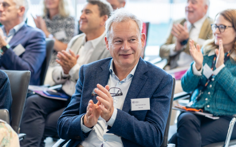 A group of people sit in rows at an event and applaud. A person in the foreground wears a blue blazer and holds their hands together while others in the background clap.