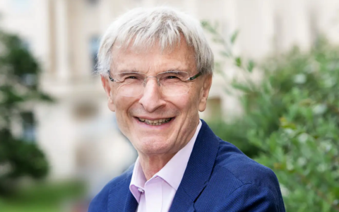 A person with short light‑grey hair wearing a blue blazer over a light pink collared shirt, photographed outdoors with greenery and a light-colored building in the background