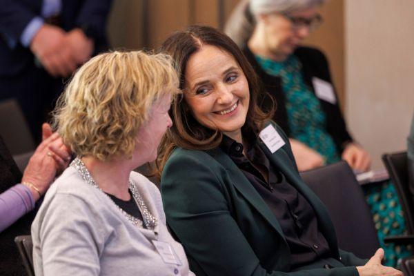 a group of people sitting in chairs at an indoor event with one person in the foreground wearing a light colored top and another person next to them wearing a dark green jacket