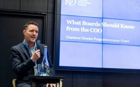 Speaker presenting at an institute of directors ireland event, standing at a podium with a microphone and water bottle, with a screen behind displaying the title what boards should know from the coo and chartered director programme event.