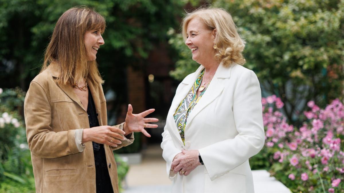 Two people standing outdoors engaged in conversation. The person on the left is wearing a tan jacket over a black top and gesturing with both hands. The person on the right is wearing a white blazer over a patterned blouse with their hands clasped in front of them. The background features green plants and pink flowers.