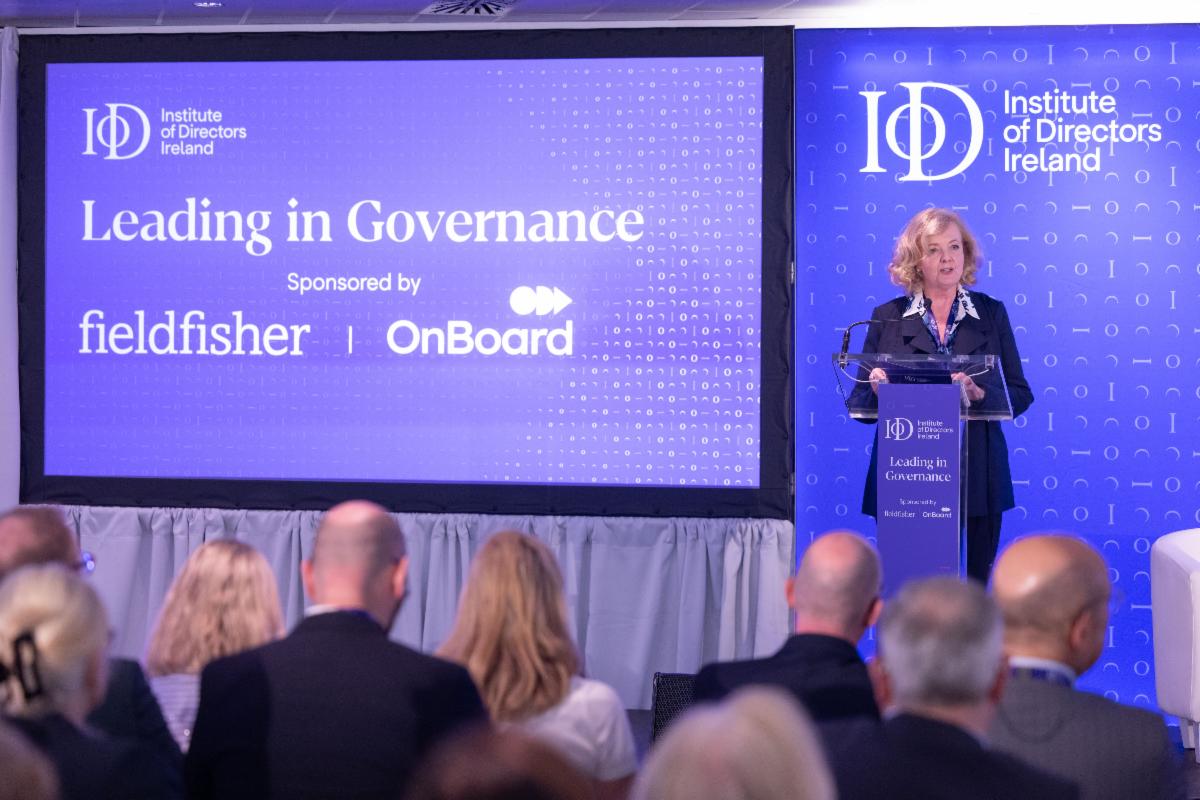 Conference setting with a speaker standing at a clear podium on the right in front of a branded backdrop for Institute of Directors Ireland. A large screen on the left displays the text Institute of Directors Ireland Leading in Governance Sponsored by fieldfisher and OnBoard. Several attendees are seated in the foreground facing the stage.