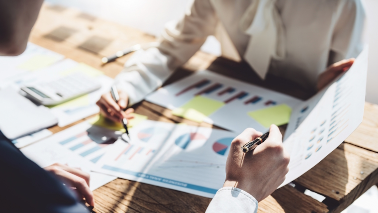 Two people seated at a wooden table reviewing printed charts and graphs with a calculator and documents suggesting a business meeting or financial planning session