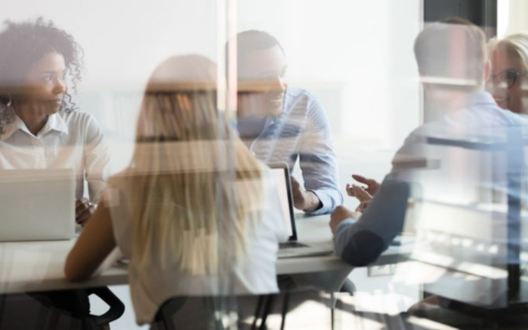 Group of people seated around a table in a modern office setting engaged in discussion. Laptops and documents are visible on the table. The scene is viewed through a glass partition creating reflections that add depth to the image. Bright natural light fills the room suggesting a collaborative work environment.