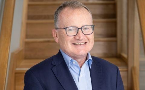 Professional headshot of a man wearing a navy blazer over a light blue shirt. He is standing indoors in front of a wooden staircase background.