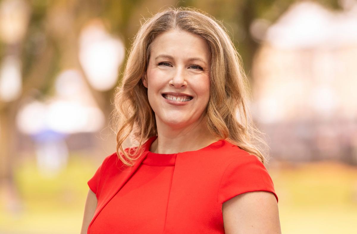 a person with long blonde hair wearing a red dress standing outdoors with a blurred background of trees and greenery