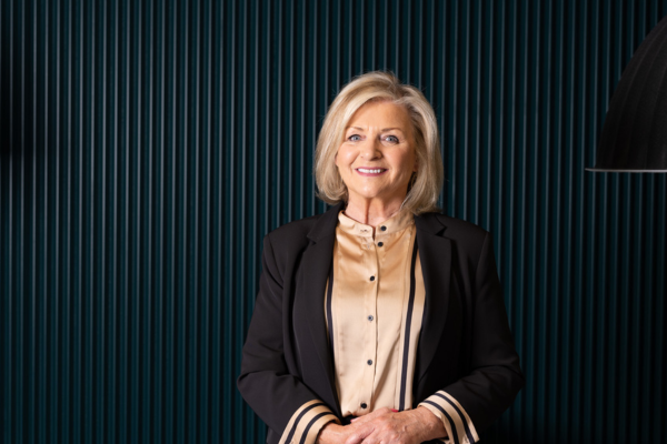 person with blond hair black jacket and gold blouse facing the camera and smiling. against dark background