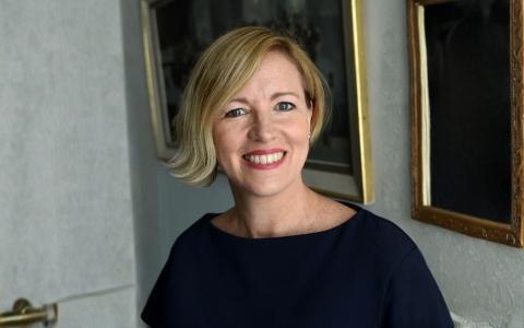 Professional headshot of an individual wearing a dark top, photographed indoors against a softly blurred background. The portrait is cropped from the shoulders up and shows the individual facing the camera, with framed artwork visible in the background. 