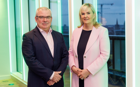 Two individuals standing side by side in a bright indoor space with large windows in the background. Their faces are blurred. The person on the left wears a dark suit with a light pink shirt and the person on the right wears a light pink coat over a black dress.