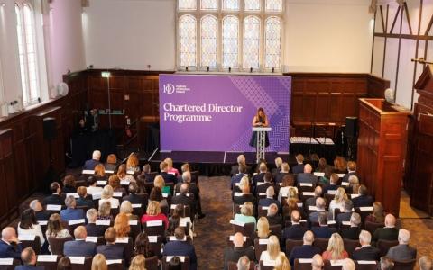 Audience seated in a large hall for an Institute of Directors Ireland Chartered Director Programme event with a speaker presenting on stage in front of a branded screen.