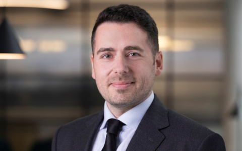 Professional portrait of a man wearing a dark suit, white shirt, and black tie. He is standing indoors with a softly lit, modern office background.