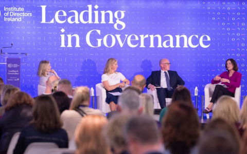 Panel discussion on stage with four individuals seated on white chairs in front of a bright blue backdrop displaying the words Leading in Governance. Each person is engaged in conversation and holding notes or gesturing. Bottles of water are placed beside the chairs. Audience members are visible in the foreground facing the panel.