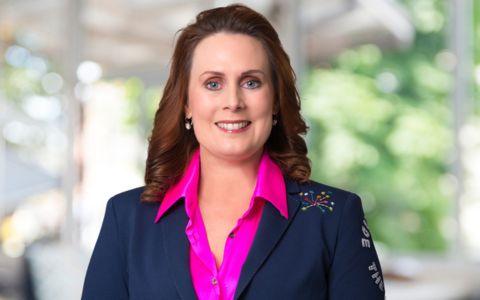 Professional portrait of a woman wearing a navy blazer with embroidered detail over a bright pink blouse. She is standing indoors with a softly lit, blurred background of greenery and architectural elements.