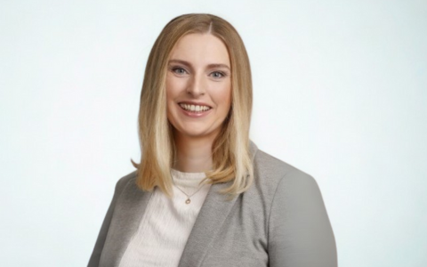 Person with shoulder length straight hair wearing a light grey blazer and white top with a small pendant necklace standing in front of a plain light background