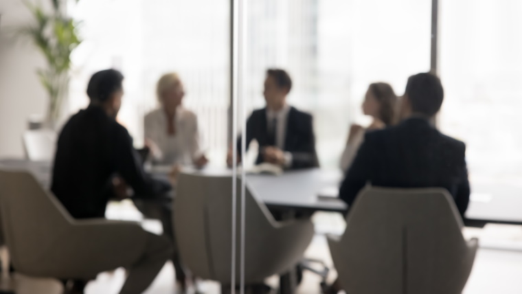 group of people in business attire seated around a board table. Image blurred behind glass