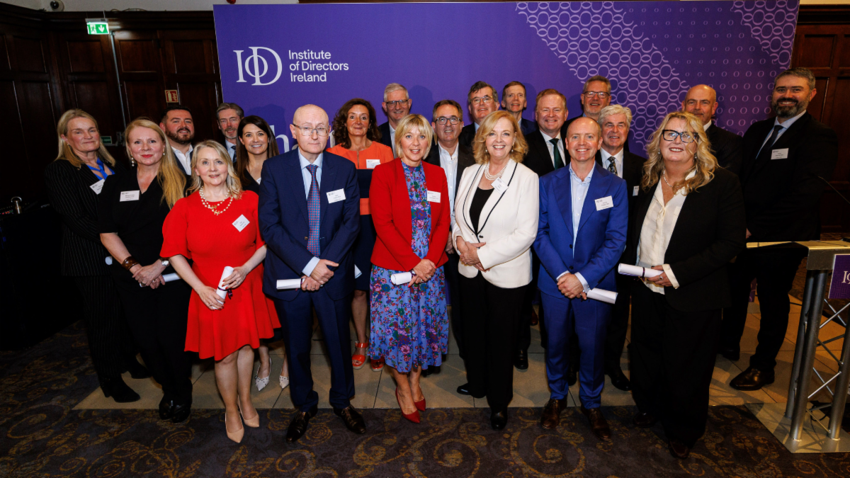 Group of people in business attire standing in front of a purple backdrop with Institute of Directors Ireland logo and hexagonal pattern some holding rolled up documents suggesting a formal event or ceremony