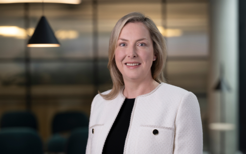 Professional headshot of a person wearing a light textured jacket over a dark top, standing in an office setting with soft lighting and blurred background details including a hanging pendant light.