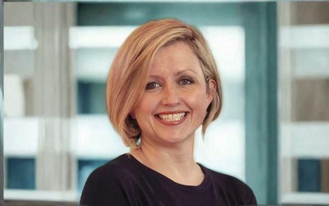 Professional portrait of a woman with short blonde hair wearing a black top in a modern office setting. Smiling to camera.