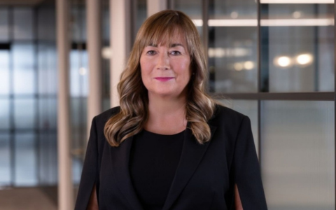 Professional headshot of a person standing in a modern office corridor. The person is wearing a black outfit with a cape-style blazer. Glass partition walls and softly lit office interiors appear in the background.
