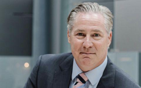 Professional portrait of a man wearing a dark suit, light blue shirt, and striped tie. He is seated indoors with a modern, softly lit office background.