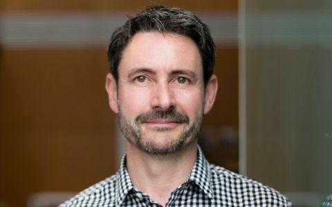 Professional headshot of a man wearing a checked shirt standing in an office environment.