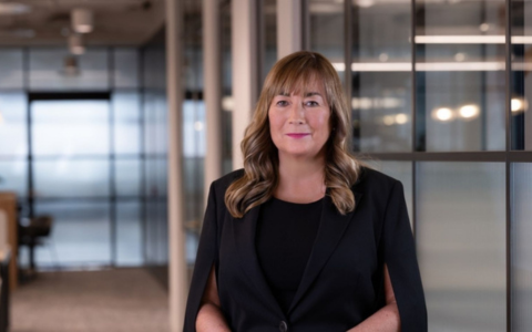 person with long brown hair and black suit facing camera in front of a blurred office background