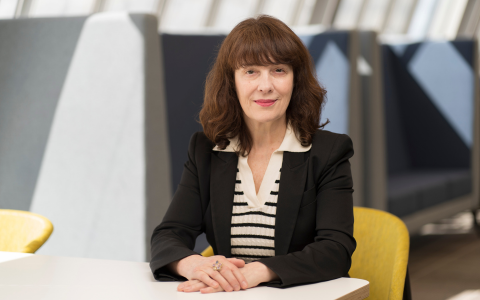 Professional portrait of a woman seated at a table wearing a black blazer and patterned top in a modern office environment.