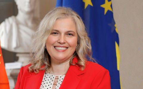 A person with shoulder‑length wavy blonde hair wearing a bright red blazer over a patterned blouse, standing indoors beside a European Union flag and a white marble statue in the background