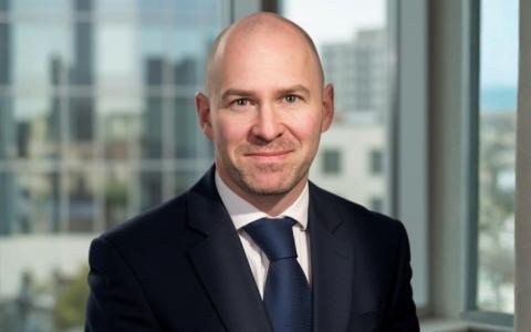 Professional headshot of an individual in business attire against a plain grey background. The portrait is cropped from the shoulders up and shows the individual facing the camera in a formal corporate style