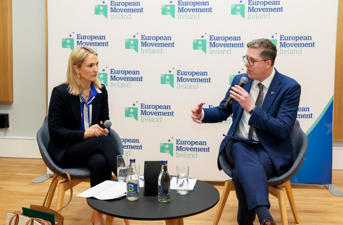 Two panelists speaking in front of a European Movement backdrop. Both seated. One holds a microphone