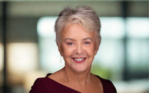 Professional portrait of a woman with short grey hair wearing a dark top. She is standing indoors with a softly blurred background of windows and greenery.