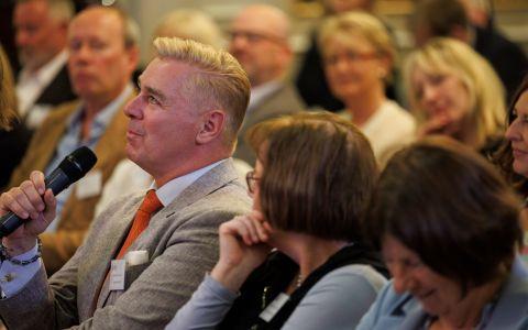 Person seated in an audience speaking into a microphone with a grey suit and red tie