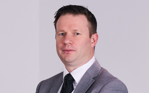 A person wearing a light grey suit jacket, white shirt, and dark tie, posed against a plain light‑coloured background with arms crossed