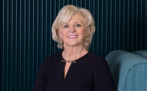 Professional headshot of a person seated against a dark blue textured backdrop. The person is wearing a dark top and a gold necklace and is photographed in an indoor setting beside a blue upholstered chair.