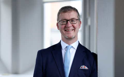 Professional headshot of a person wearing glasses, a dark suit jacket, white shirt, and light blue tie, standing indoors beside a wall in a softly lit office setting.