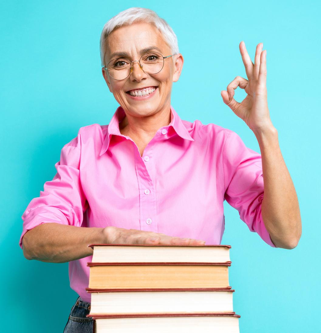 Older woman in a pink shirt with a pile of books giving the ok sign