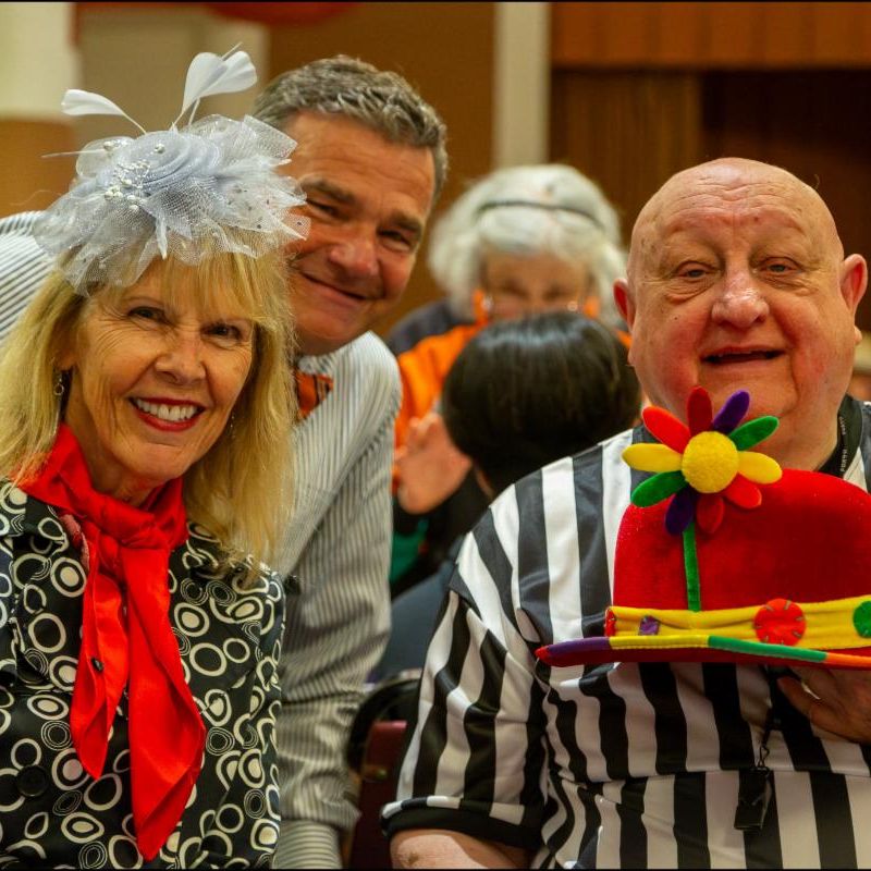Three smiling people at an indoor party, one woman in a decorative hat and red scarf, a man in a striped shirt wearing a festive holiday hat, with a crowd in the background.