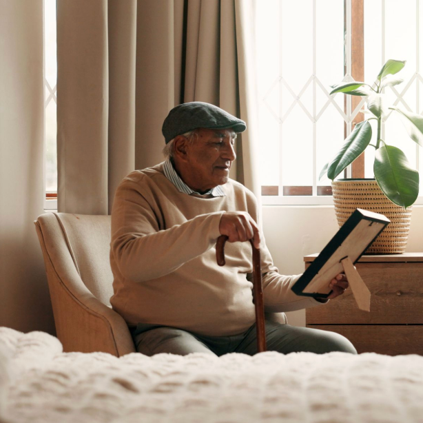An older man wearing a cap sits in an armchair by a sunlit window, holding a cane and a book.