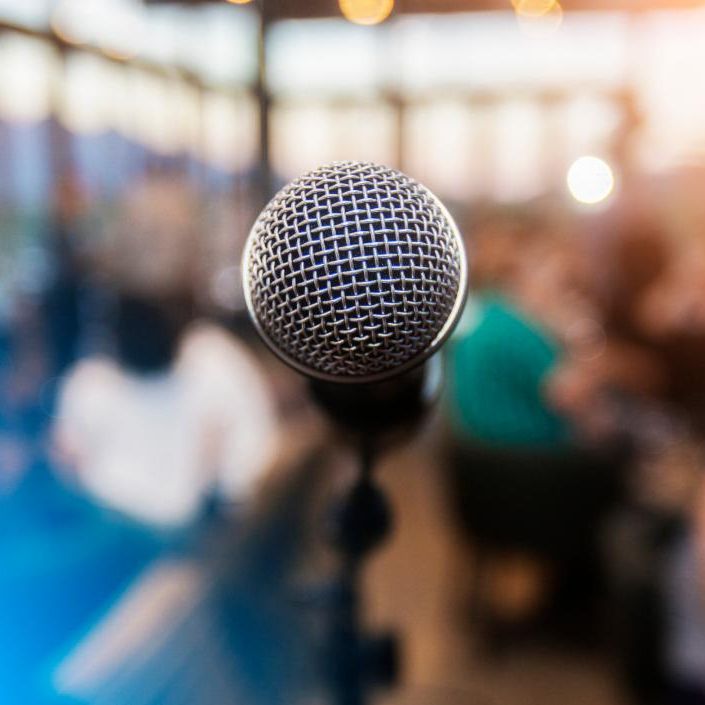Close-up of a microphone on a stand, with a blurred audience in the background.