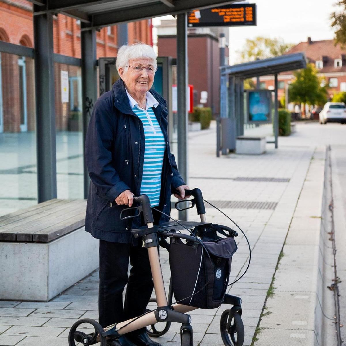 An older woman with a rollator walker stands on a city sidewalk near a bus stop.