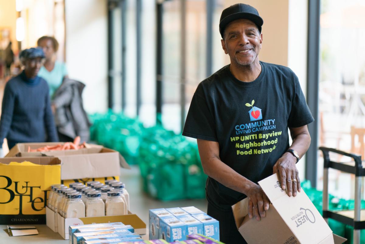 A smiling man in a CLC Bayview Neighborhood Food Network T-Shirt packs a box of groceries at a food distribution table stocked with milk, produce and snacks.