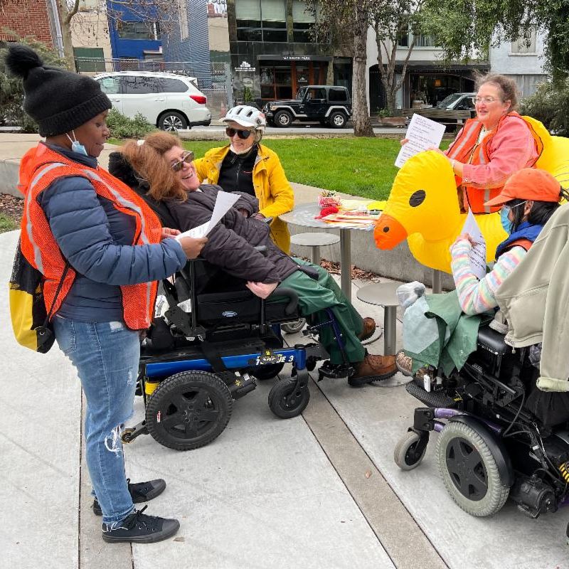 A small group gathered outdoors around a table, including people using power wheelchairs, one wearing an orange safety vest, chatting near a large yellow duck decoration.