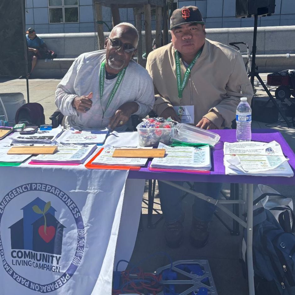 Paul and Mark wearing lanyards stand behind an outdoor table with colorful flyers and a Community Living Campaign banner.